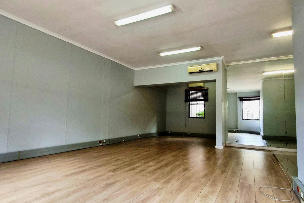 An empty office space featuring laminate flooring, fluorescent lighting, a wall-mounted air conditioning unit, and a large mirrored wall section.
