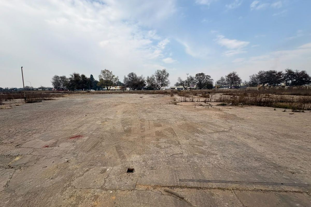 A wide-angle view of a vacant commercial land plot featuring a large, weathered concrete surface with visible cracks and surrounding vegetation.