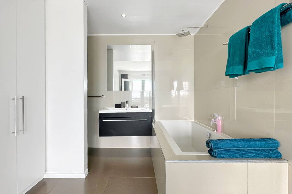 Modern bathroom featuring a wall-mounted vanity, built-in bathtub with overhead shower, and integrated storage cabinetry with neutral tiled finishes.