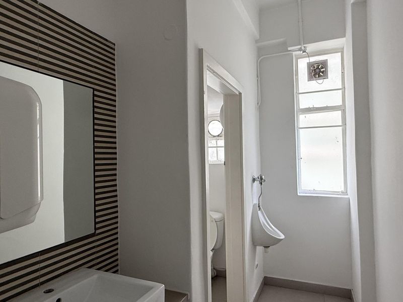 A functional office bathroom featuring a wall-mounted basin, a wall-mounted urinal, and a tiled feature wall with a mirror.