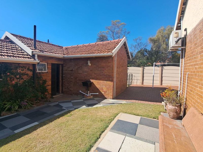 A well-maintained rear exterior view featuring a brick facade, tiled roof, paved patio area, and a small garden space.