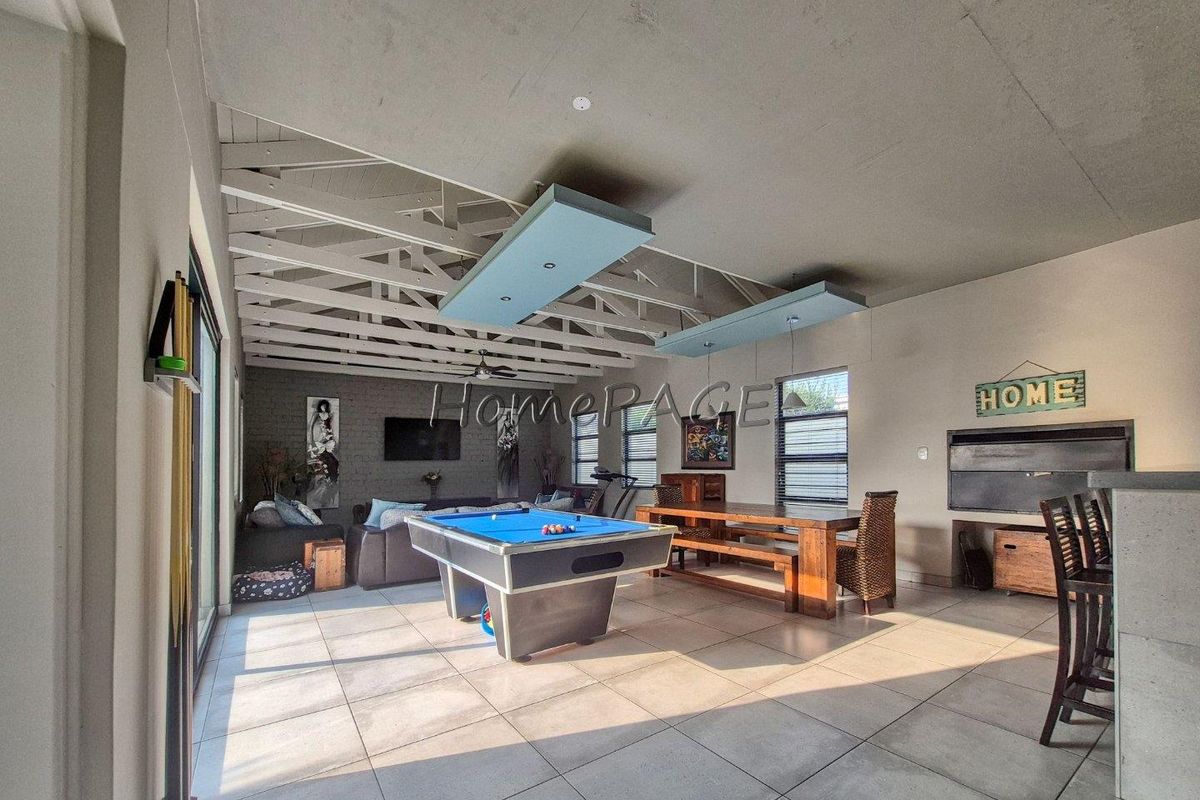 Spacious living area featuring exposed timber roof trusses, tiled flooring, and a built-in braai, opening out through sliding glass doors.