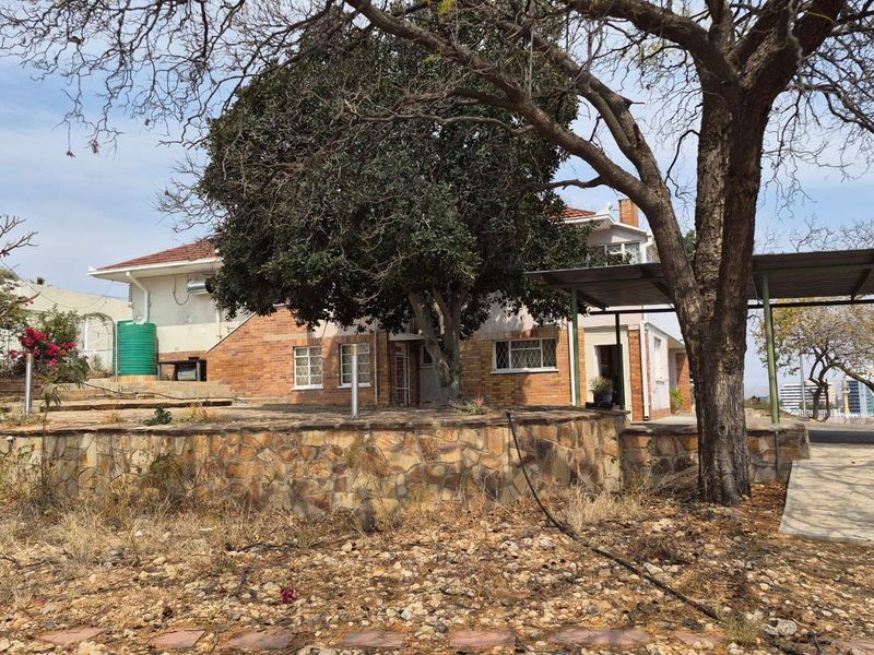 Rear view of the property featuring a brick facade, stone retaining wall, and a carport, set within a large garden area.