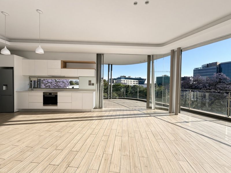 Modern open-plan living area featuring a sleek white kitchen, wood-look tiled flooring, and expansive floor-to-ceiling glass doors opening onto a spacious balcony with city views.