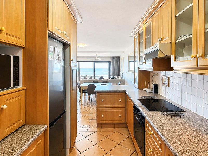 Galley-style kitchen featuring light wood cabinetry, laminate countertops, and integrated appliances, flowing into an open-plan living area with ocean views.