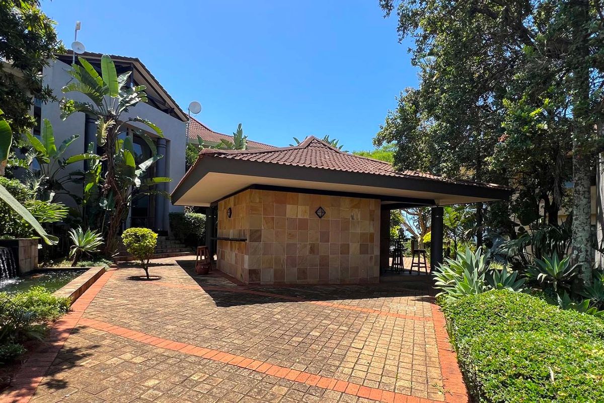 A well-maintained outdoor entertainment area featuring a stone-clad bar structure, paved patio, and a tranquil water feature set within a lush garden.