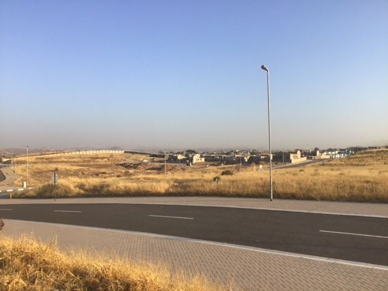 A view of the paved street and surrounding vacant residential land with established infrastructure including street lighting and boundary walls.