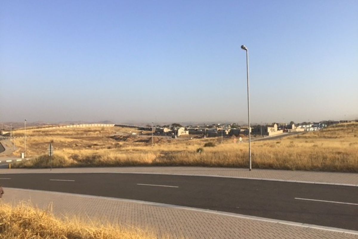A view of the paved street and surrounding vacant residential land with established infrastructure including street lighting and boundary walls.
