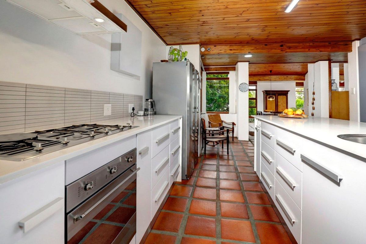 Modern kitchen featuring white cabinetry, stone countertops, integrated gas hob, and oven, set against a rustic terracotta tiled floor and timber-paneled ceiling.