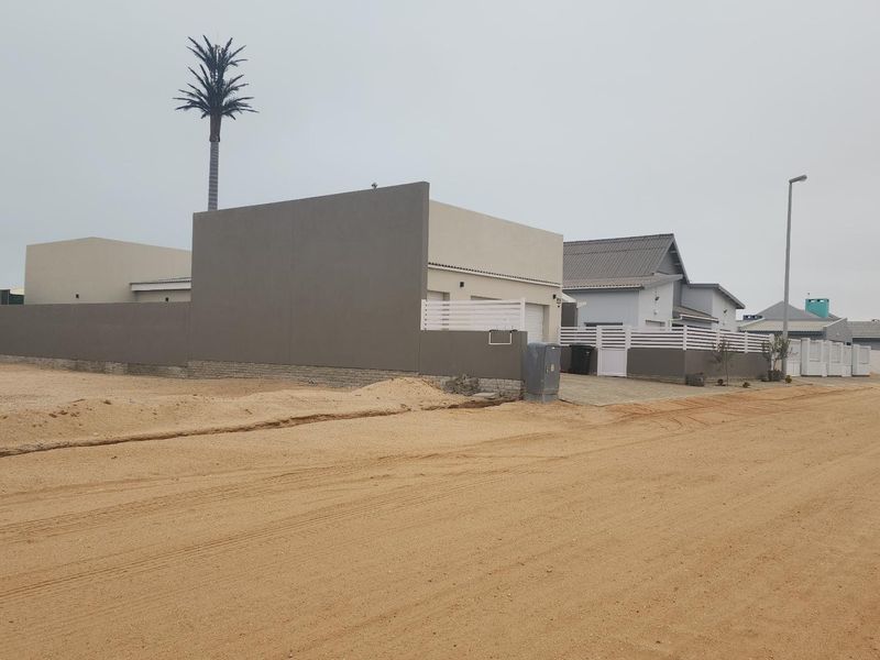 Street view of a residential property featuring a modern boundary wall, paved driveway, and gate, situated along an unpaved sandy road.