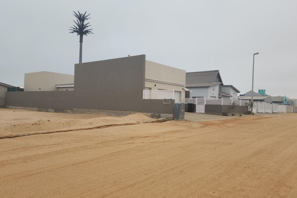 Street view of a residential property featuring a modern boundary wall, paved driveway, and gate, situated along an unpaved sandy road.