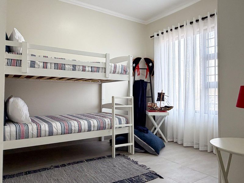A well-maintained bedroom featuring neutral tiled flooring, cove cornicing, and large windows dressed with sheer curtains.