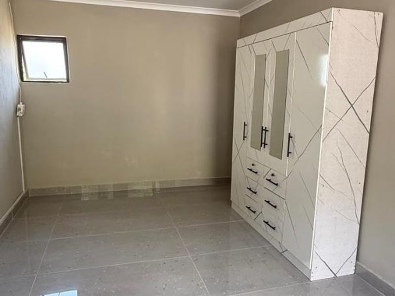 A bright, empty bedroom featuring large format grey floor tiles, neutral wall paint, and a modern built-in wardrobe with a marble-effect finish.