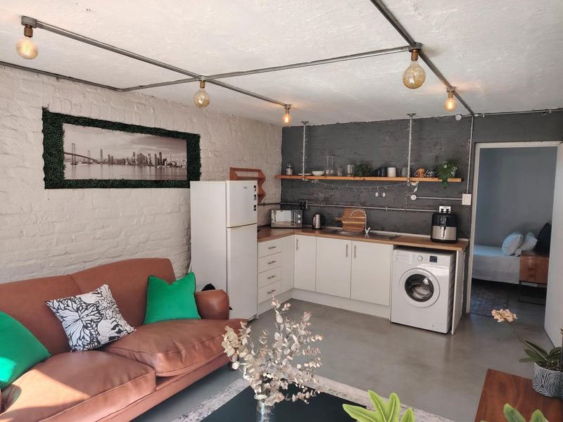 An open-plan living and kitchen area featuring industrial-style exposed conduit lighting, white cabinetry, wooden countertops, and textured exposed brick walls.