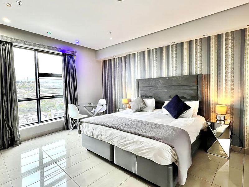 A modern, well-lit bedroom featuring polished tiled flooring, a large window with city views, and a feature wall with built-in wardrobe storage.