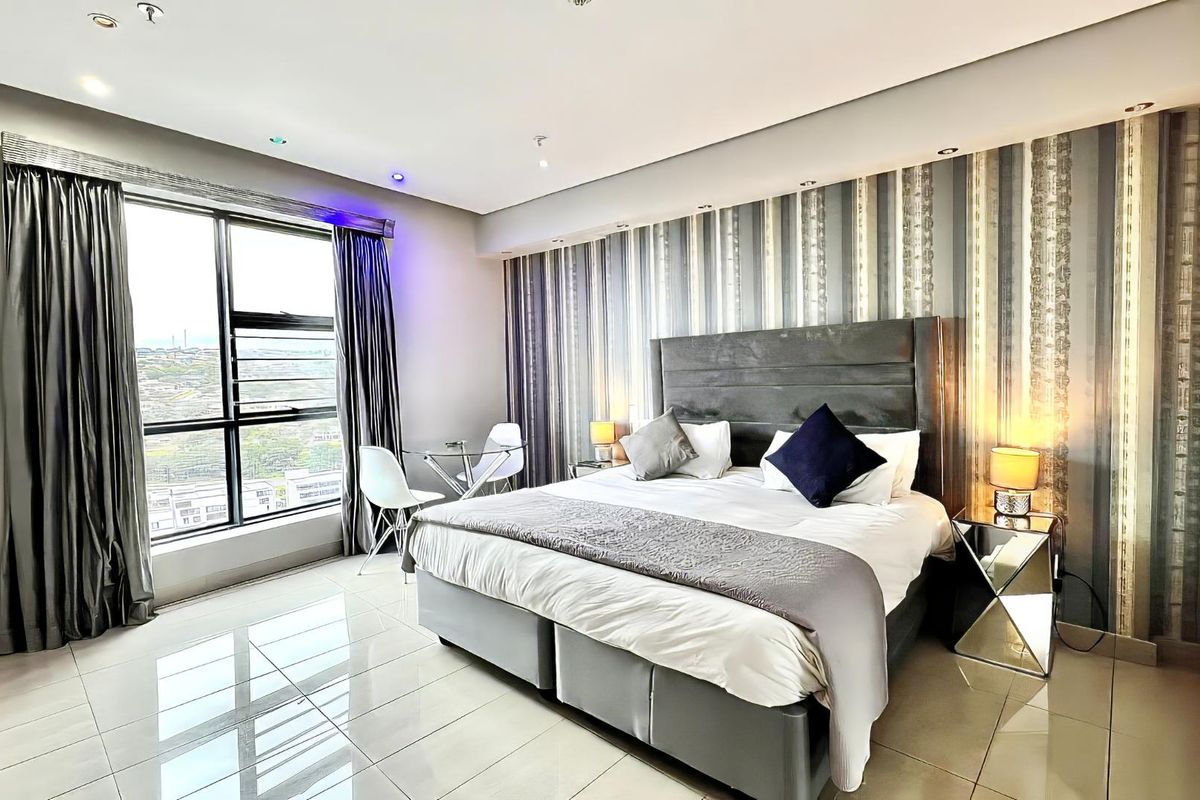 A modern, well-lit bedroom featuring polished tiled flooring, a large window with city views, and a feature wall with built-in wardrobe storage.