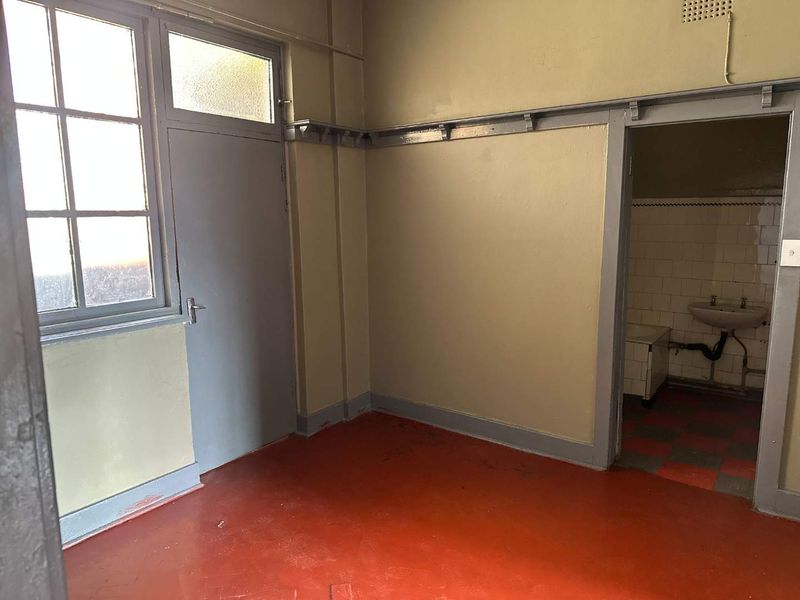 An empty utility or service room featuring a multi-pane window, painted walls, and an adjacent small wash area with a wall-mounted basin.