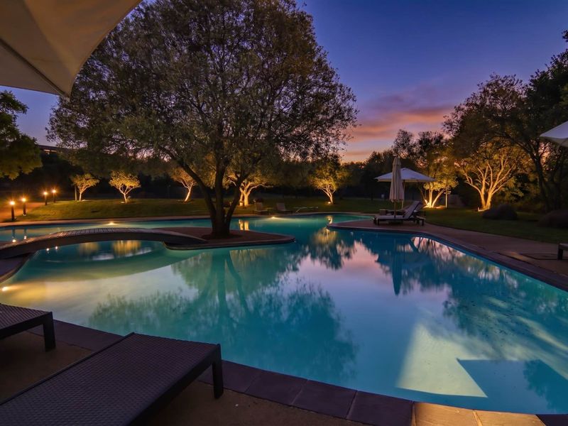 A beautifully illuminated swimming pool area at dusk, featuring a pedestrian bridge, mature trees, and well-maintained landscaping.
