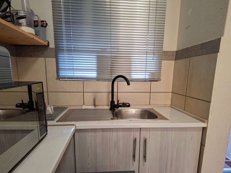 A functional kitchen area featuring a stainless steel sink, black faucet, tiled backsplash, and light wood-look cabinetry with laminate countertops.