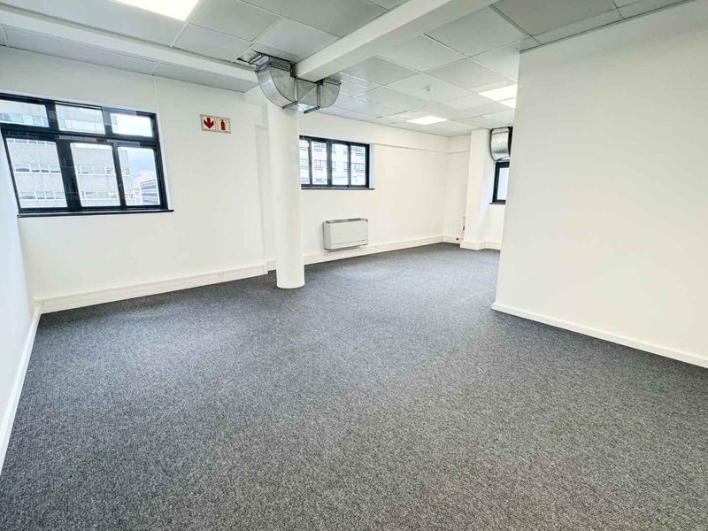 A bright, open-plan office space featuring neutral carpeting, suspended ceilings with recessed lighting, and large windows providing ample natural light.
