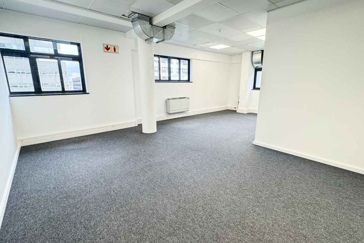 A bright, open-plan office space featuring neutral carpeting, suspended ceilings with recessed lighting, and large windows providing ample natural light.