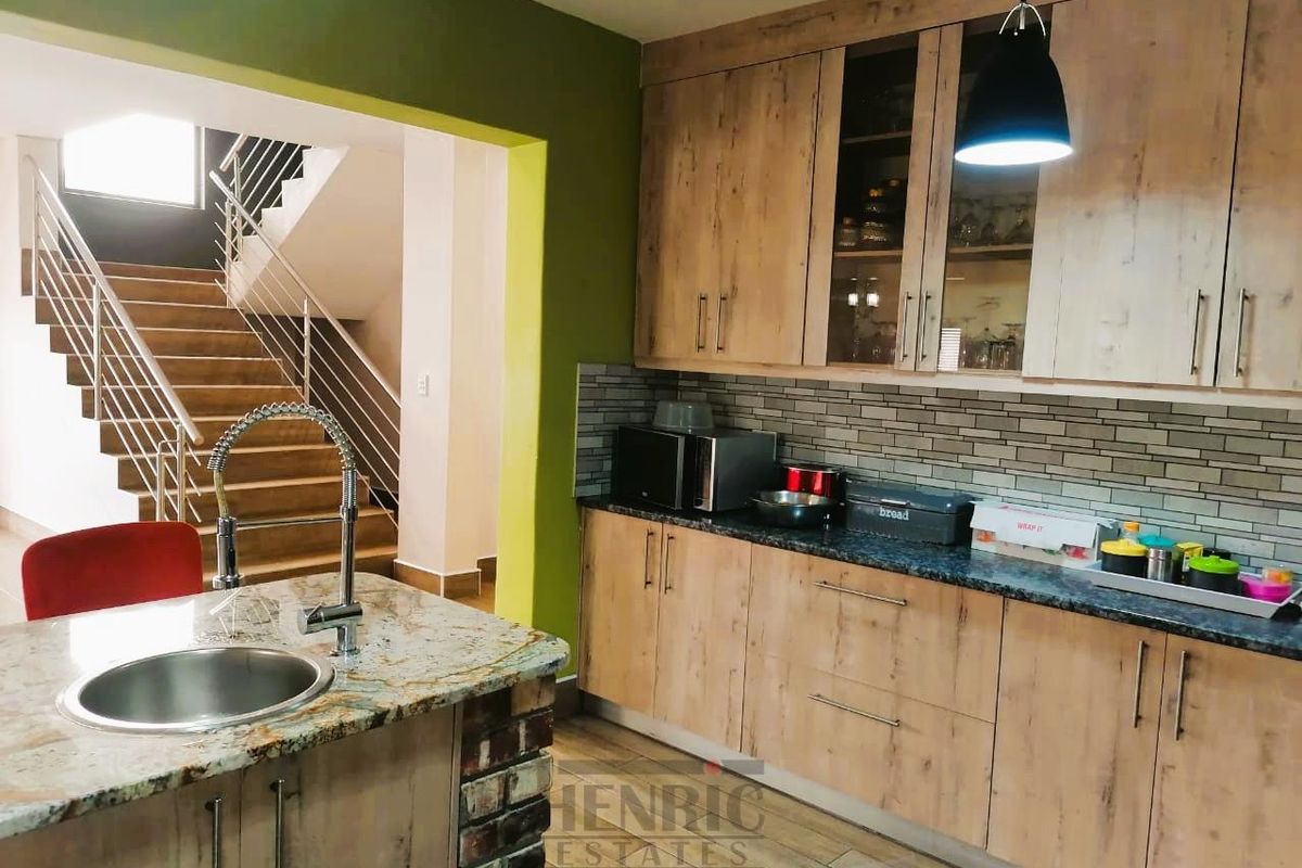 Modern kitchen featuring wood-finish cabinetry, granite countertops, a central island with a sink, and an adjacent staircase leading to the upper level.