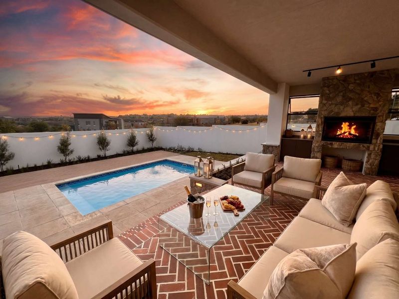 A beautifully appointed covered patio featuring a stone-clad built-in fireplace, herringbone brick flooring, and direct access to a private swimming pool at sunset.