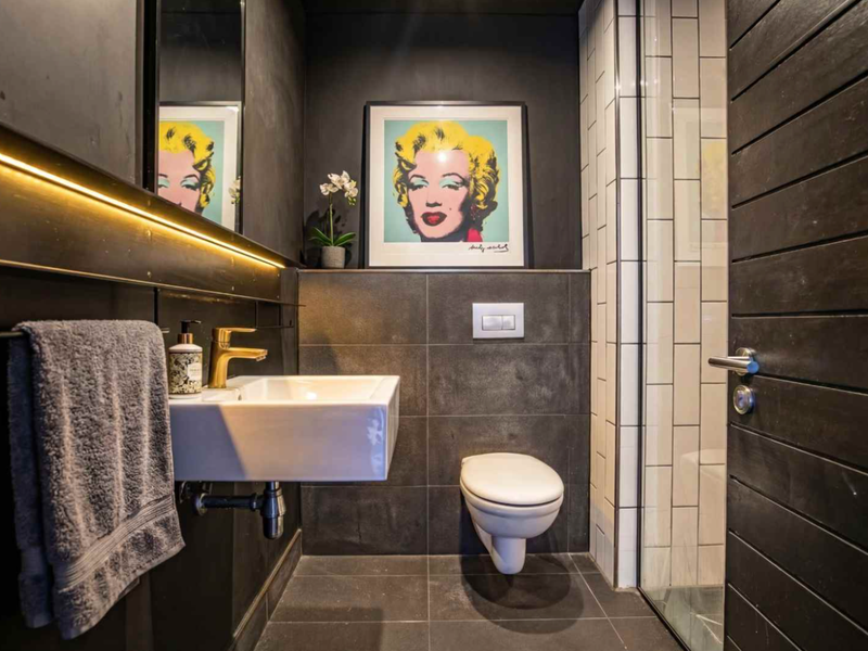 Modern bathroom featuring dark tiled finishes, wall-mounted basin with brass tap, wall-hung toilet with concealed cistern, and integrated LED lighting.
