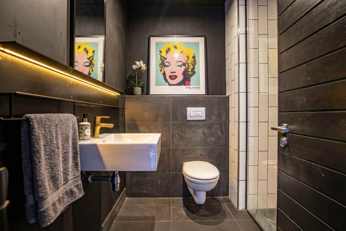Modern bathroom featuring dark tiled finishes, wall-mounted basin with brass tap, wall-hung toilet with concealed cistern, and integrated LED lighting.
