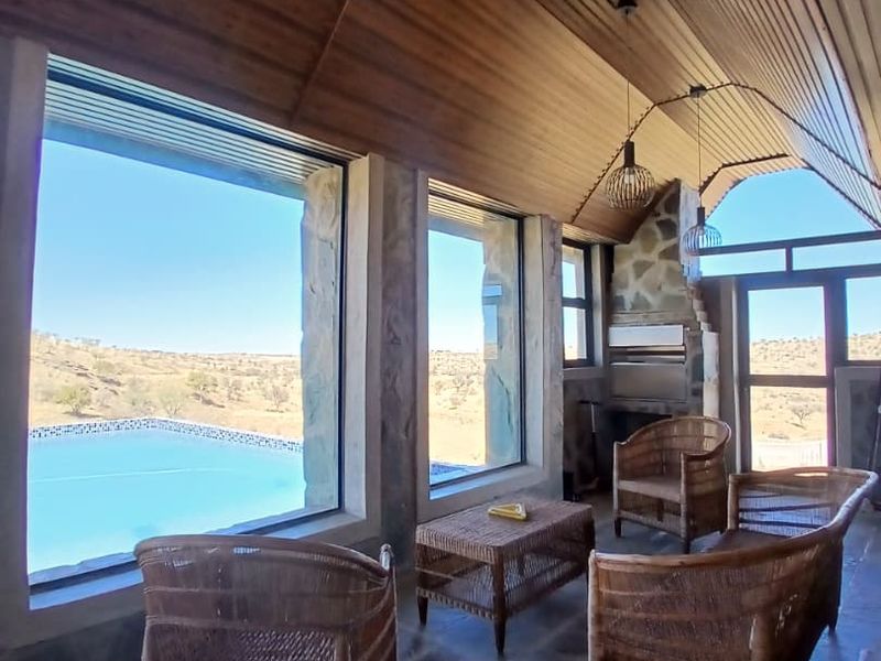 Covered patio area featuring a vaulted timber ceiling, stone accents, built-in braai, and expansive windows overlooking the swimming pool and landscape.