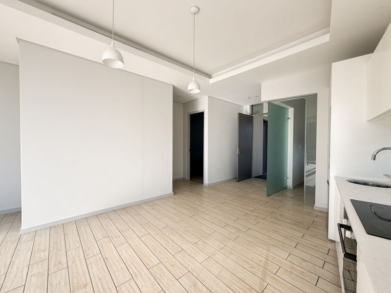 Modern open-plan living area featuring light wood-look tiled flooring, recessed ceiling details, pendant lighting, and a sleek kitchen with integrated appliances.