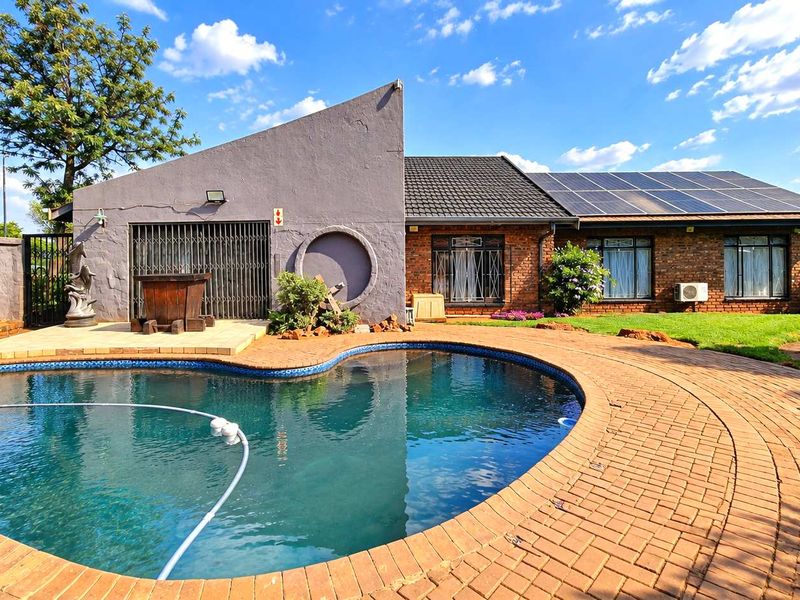 A well-maintained backyard featuring a curved swimming pool, brick paved surround, and a brick-faced house with solar panels installed on the roof.