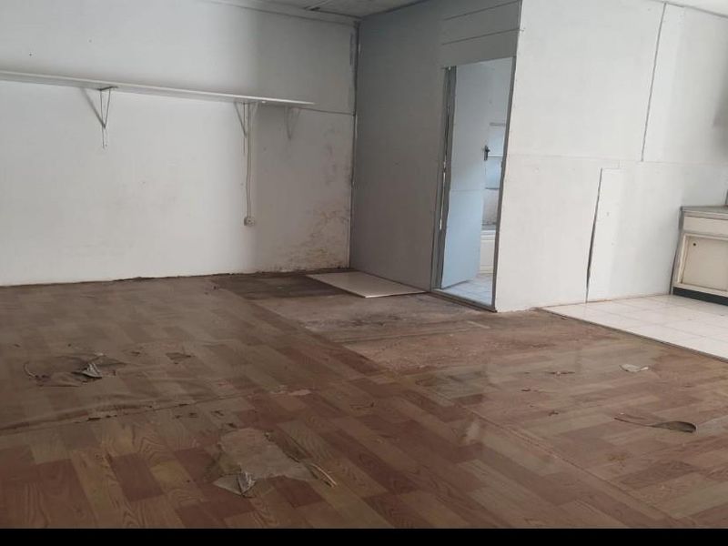An empty open-plan living and kitchen area requiring significant renovation, featuring damaged flooring, wall staining, and dated built-in cabinetry.