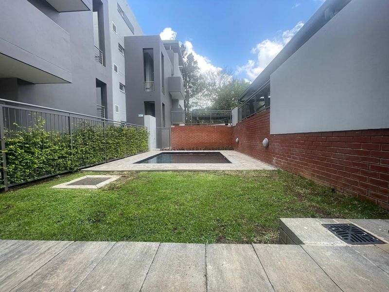A private outdoor pool area featuring a small swimming pool, paved surround, lawn, and secure perimeter fencing within an apartment complex setting.