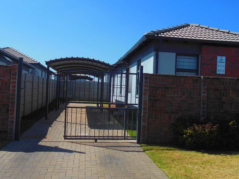 Front exterior view of the property featuring a paved driveway, covered carport, and secure metal gate with brick boundary walling.