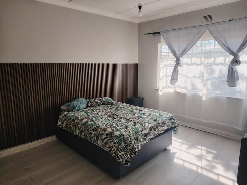 A bedroom featuring modern slatted wood wall paneling, light laminate flooring, and natural light through a window with sheer curtains.