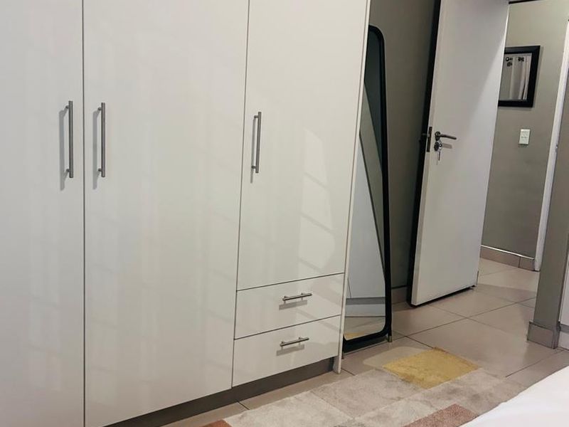 A bedroom featuring a modern white built-in wardrobe with sleek handles and neutral tiled flooring, leading into an adjacent hallway.