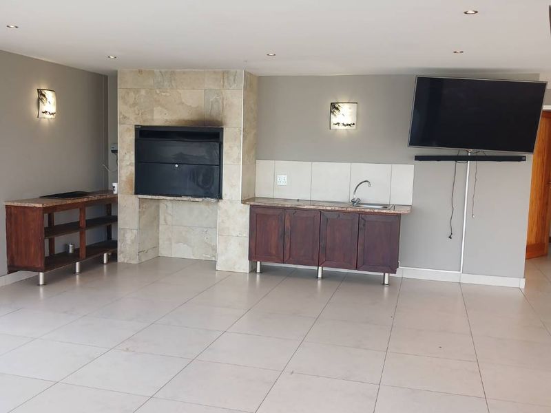 Spacious covered patio featuring a built-in braai with stone cladding, base cabinetry with a sink, and neutral tiled flooring.
