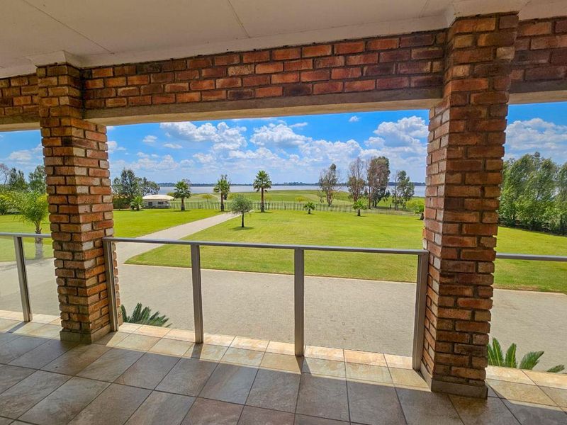 Covered patio featuring exposed brick pillars, tiled flooring, and a glass balustrade, offering expansive views over the landscaped garden and waterfront.