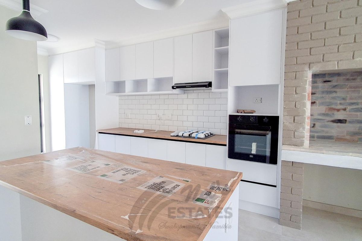 Modern kitchen featuring white cabinetry, subway tile backsplash, built-in oven, hob, and a central island, complemented by a rustic brick fireplace feature.