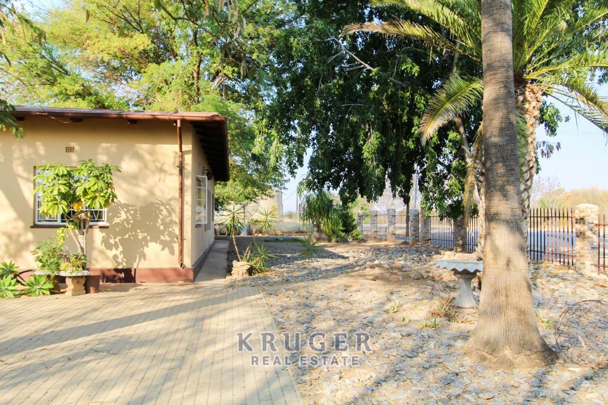 A view of the rear exterior area featuring a paved walkway, mature trees, and metal perimeter fencing.