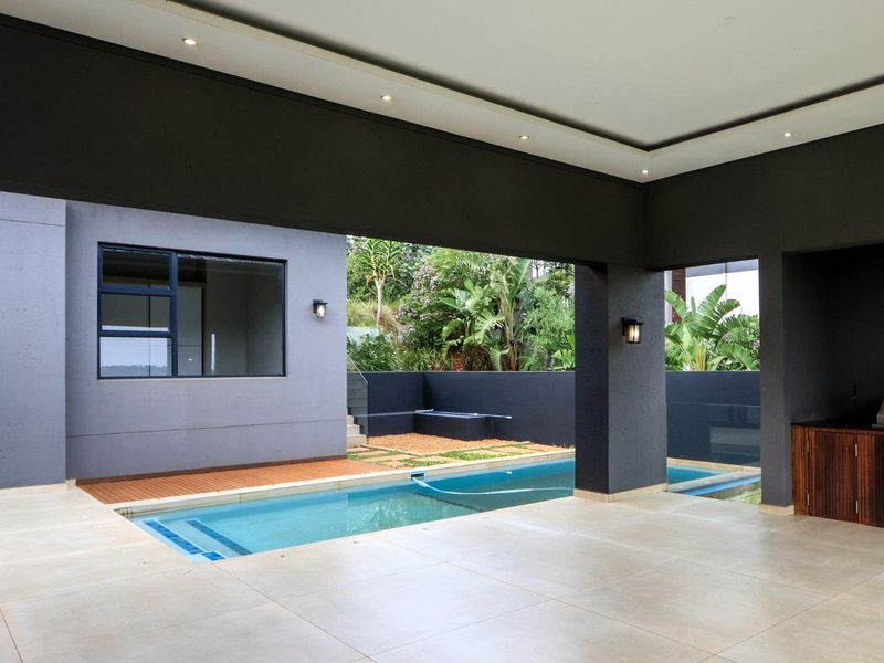 A modern covered patio featuring a built-in braai, tiled flooring, and recessed lighting, overlooking a private swimming pool and landscaped garden area.