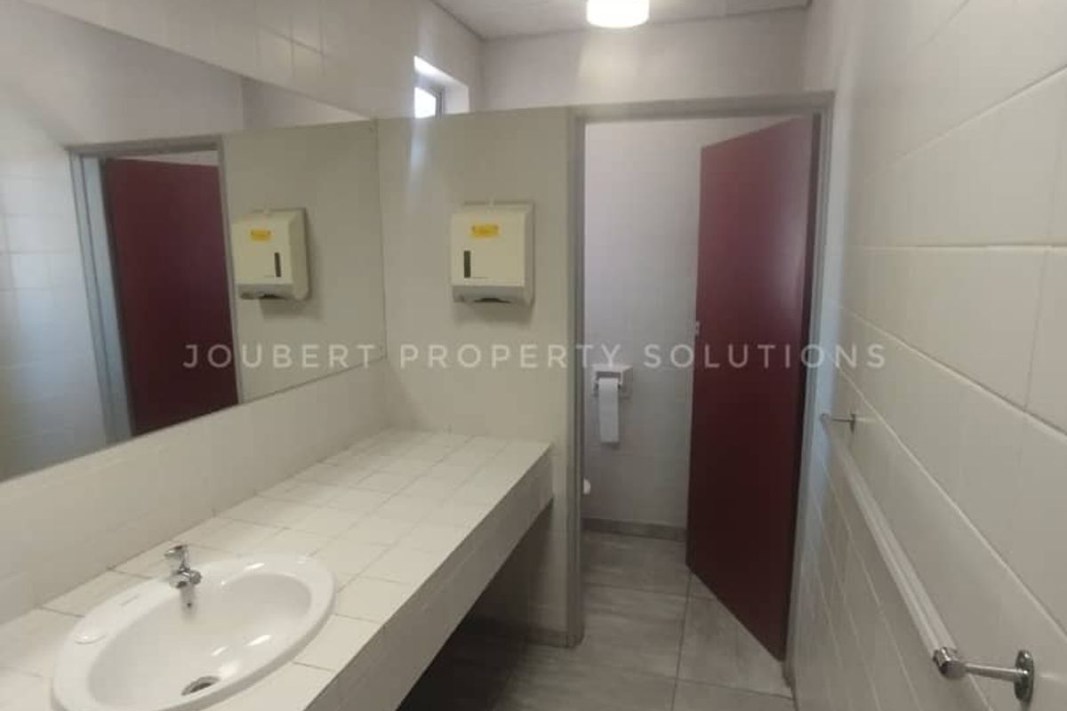 A functional commercial restroom featuring a tiled vanity countertop, integrated basin, large wall mirror, and tiled flooring.