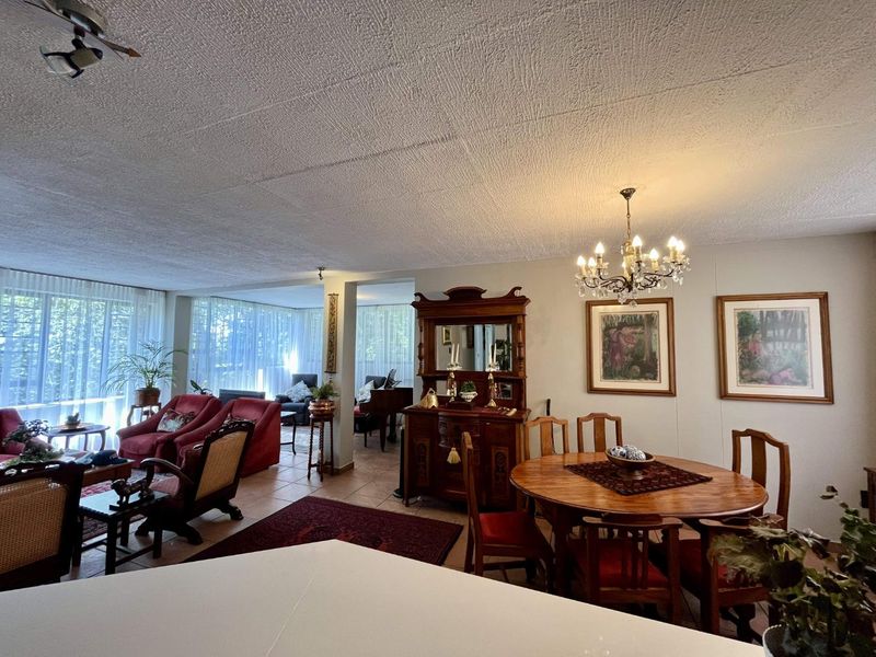 Spacious open-plan living and dining area featuring tiled flooring, large windows with natural light, and a classic chandelier.