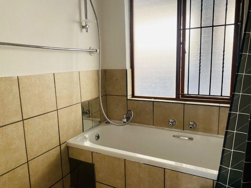 A functional bathroom featuring a built-in bathtub with a handheld shower attachment, tiled wall surrounds, and a frosted glass window with security bars.