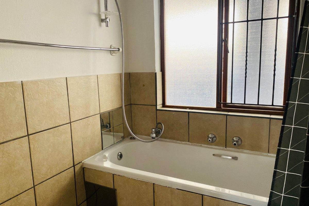A functional bathroom featuring a built-in bathtub with a handheld shower attachment, tiled wall surrounds, and a frosted glass window with security bars.