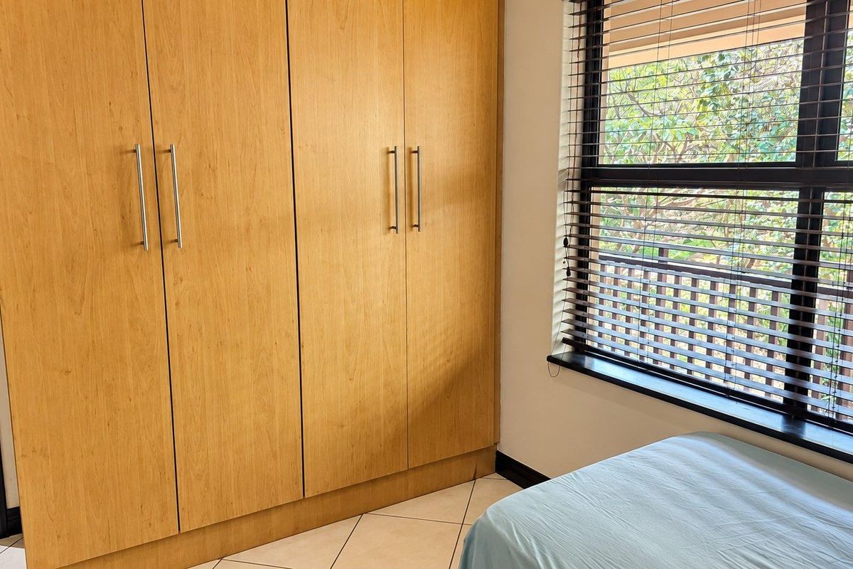 A bedroom featuring light wood built-in cupboards, tiled flooring, and a window fitted with venetian blinds.
