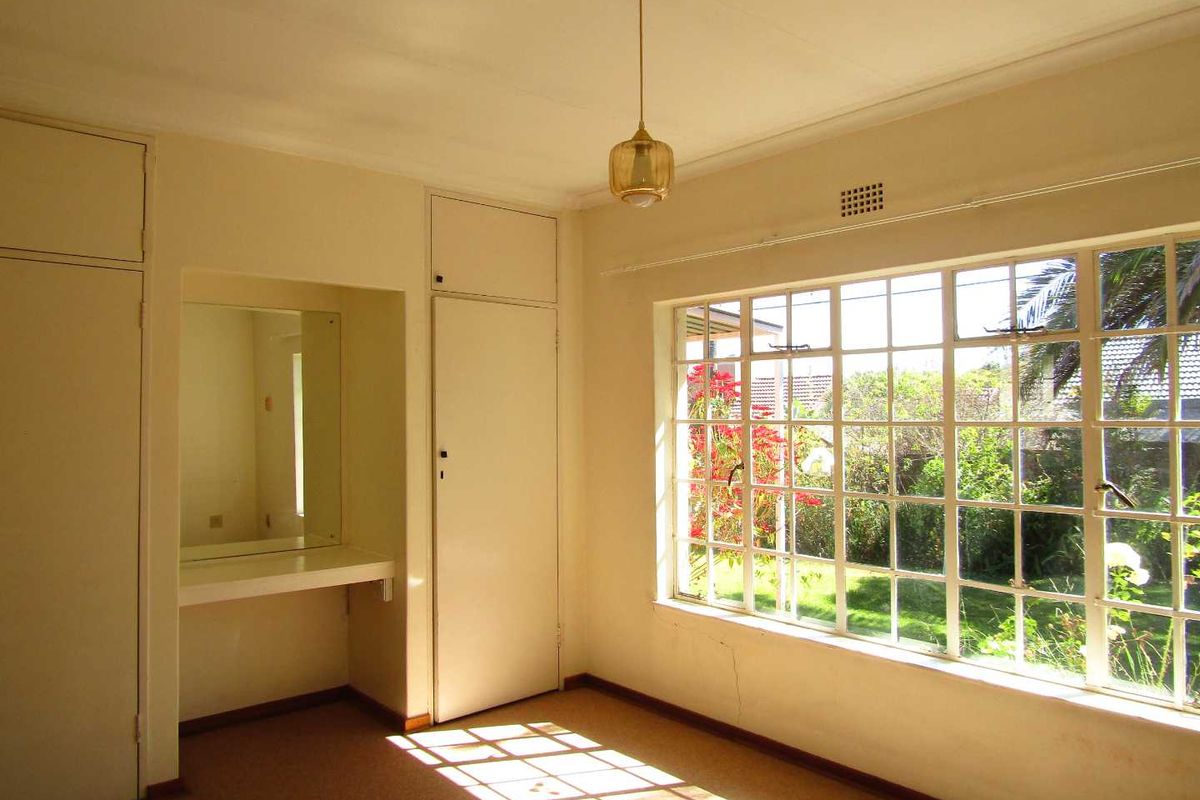 A bright, empty bedroom featuring built-in cupboards, a vanity area, and large steel-framed windows overlooking the garden.