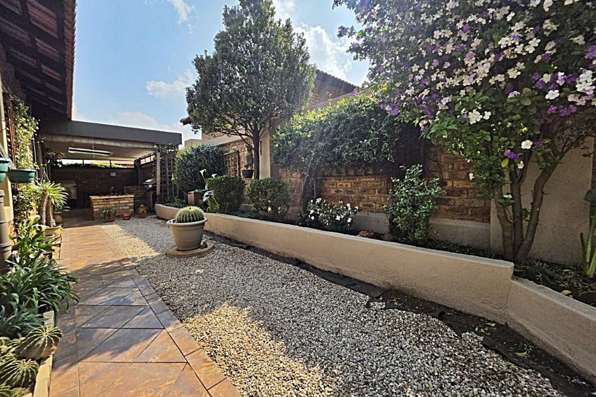 A well-maintained rear garden featuring a paved walkway, gravel landscaping, established trees, and a covered patio area with brick walling.