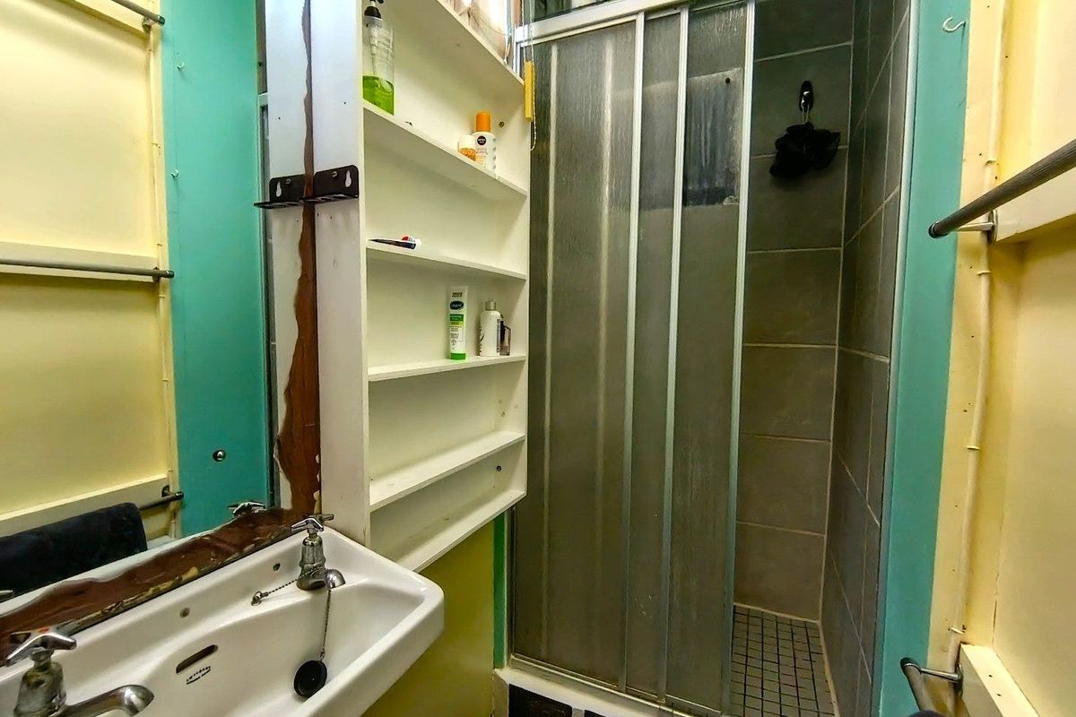 A dated bathroom featuring a pedestal basin, built-in shelving, and a shower enclosure with sliding doors, requiring cosmetic updates and repairs.